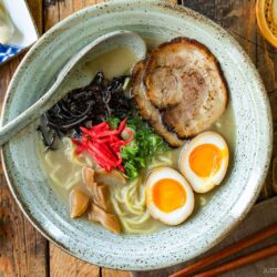 A rustic bowl containing noodles with tonkotsu broth, topped with chashu, ramen eggs, menma, kikurage, ginger, and chopped green onion. On the table, a plate of crispy gyoza and a glass of mugicha are served together.