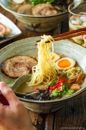 A rustic bowl containing noodles with tonkotsu broth, topped with chashu, ramen eggs, menma, kikurage, ginger, and chopped green onion. On the table, a plate of crispy gyoza and a glass of mugicha are served together.