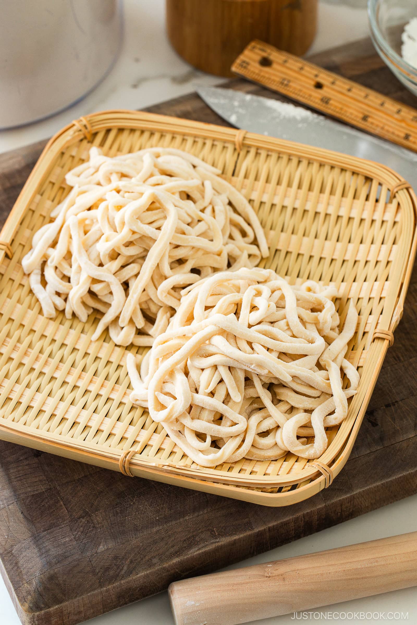 Two bundles of freshly made, uncooked udon noodles rest on a bamboo tray, placed atop a wooden cutting board with a ruler, rolling pin, and kitchen knife in the background.