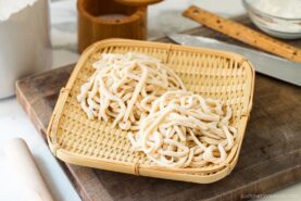 Two bundles of freshly made, uncooked udon noodles rest on a bamboo tray, placed atop a wooden cutting board with a ruler, rolling pin, and kitchen knife in the background.