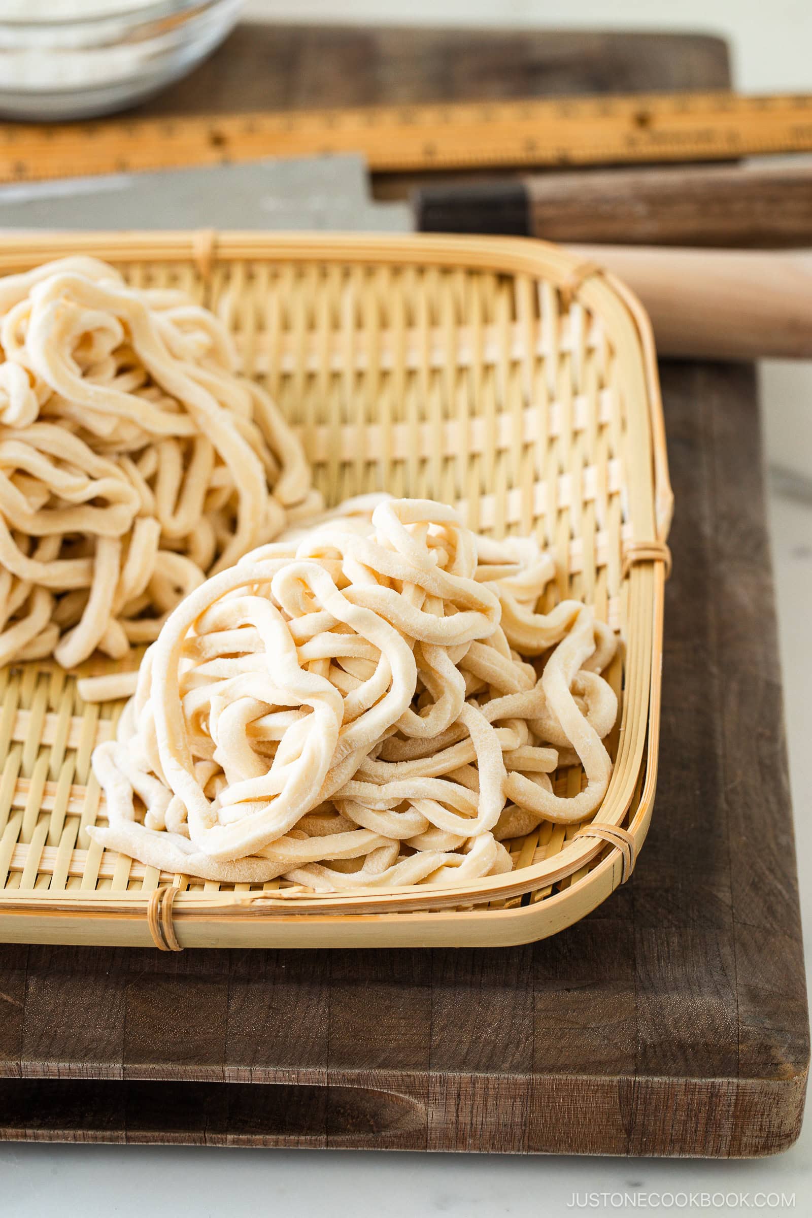 Two bundles of freshly made, uncooked udon noodles rest on a bamboo tray, placed atop a wooden cutting board with a ruler, rolling pin, and kitchen knife in the background.