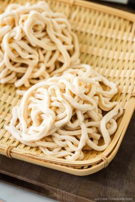 Two bundles of freshly made, uncooked udon noodles rest on a bamboo tray, placed atop a wooden cutting board with a ruler, rolling pin, and kitchen knife in the background.