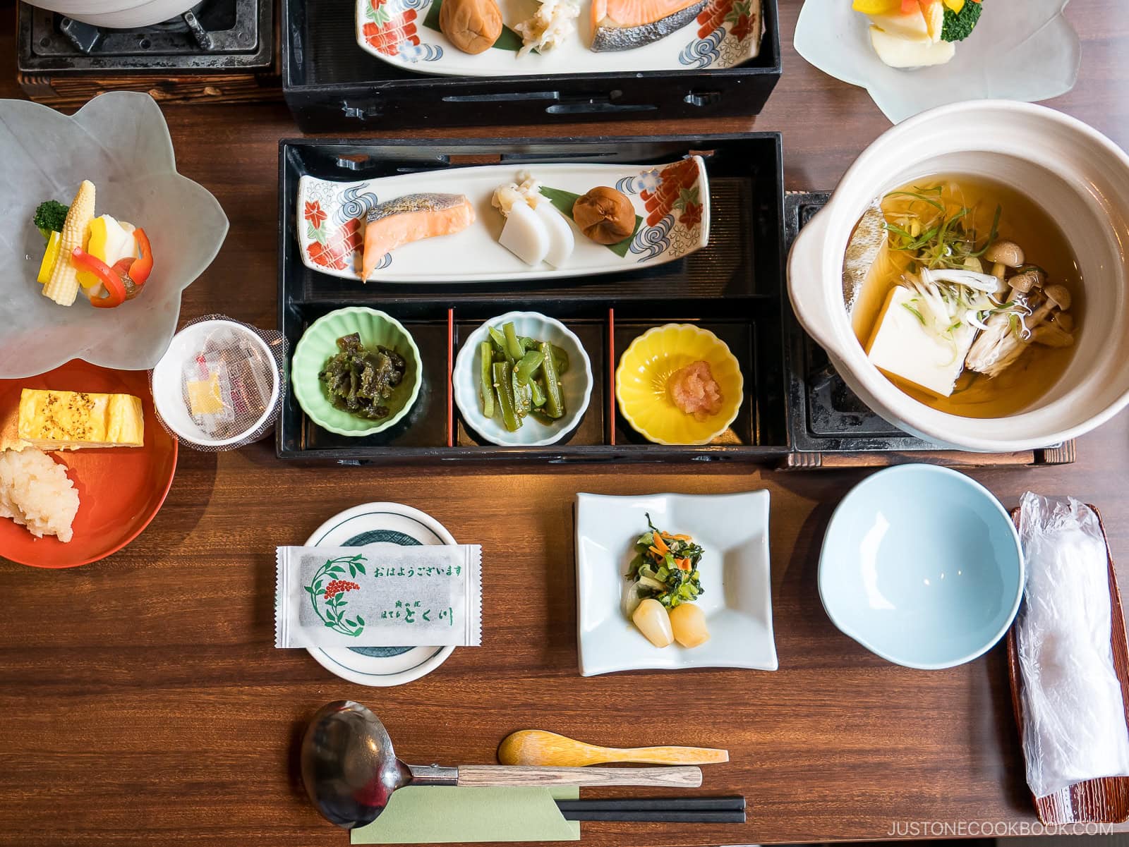 A traditional Japanese breakfast spread with grilled fish, tofu soup, assorted pickles, vegetables, tamagoyaki, rice, and small side dishes, neatly arranged on a wooden table with chopsticks and utensils.