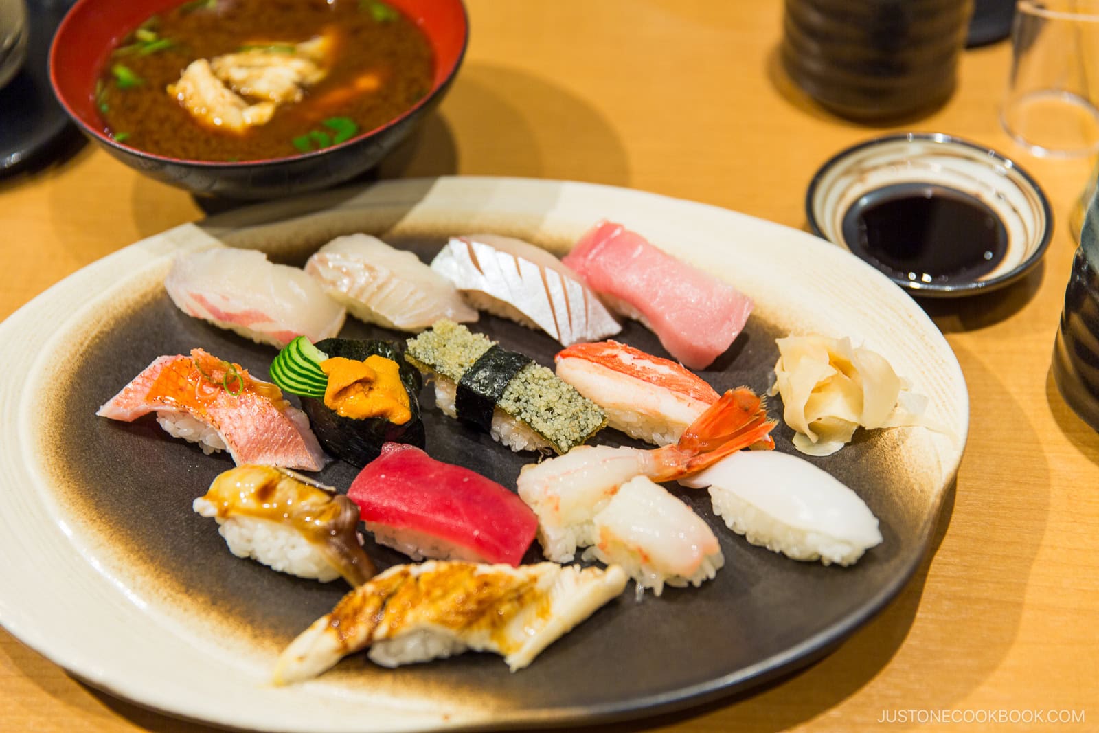 A plate of assorted sushi nigiri with various toppings, a small mound of wasabi, pickled ginger, a bowl of soup, a dish of soy sauce, and a drinking glass on a wooden table.
