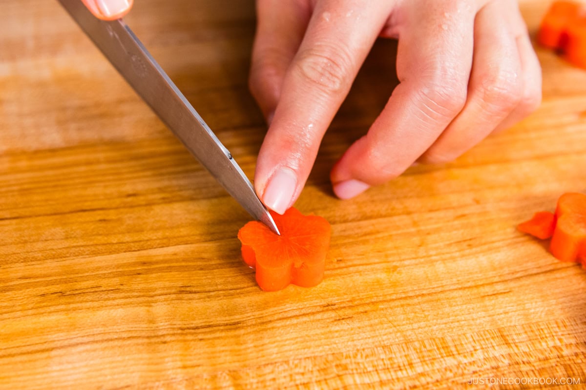 A hand uses a knife to slice a carrot piece shaped like a flower on a wooden cutting board.
