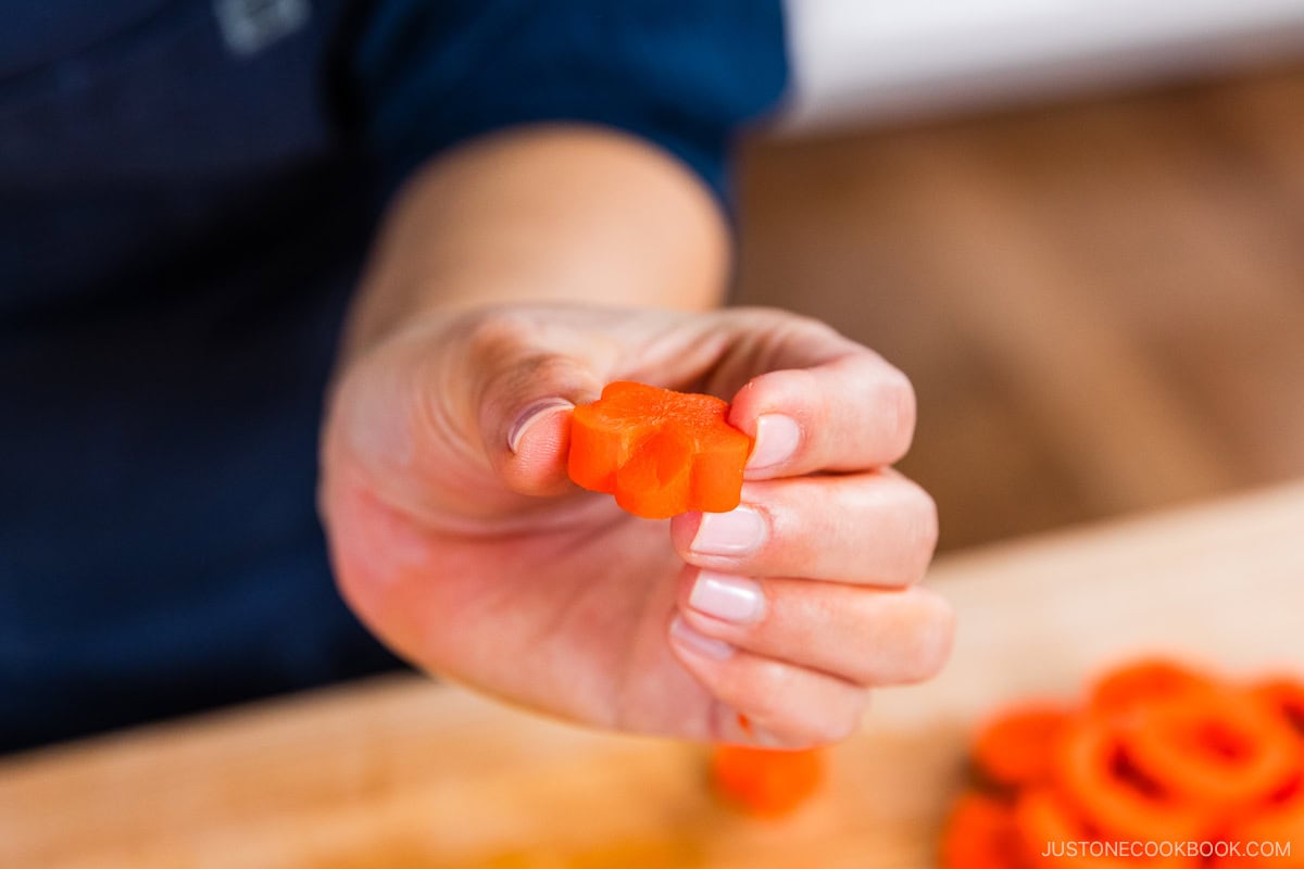 A hand holds up a slice of carrot cut into a flower shape, with more shaped carrot slices on a wooden surface in the background.