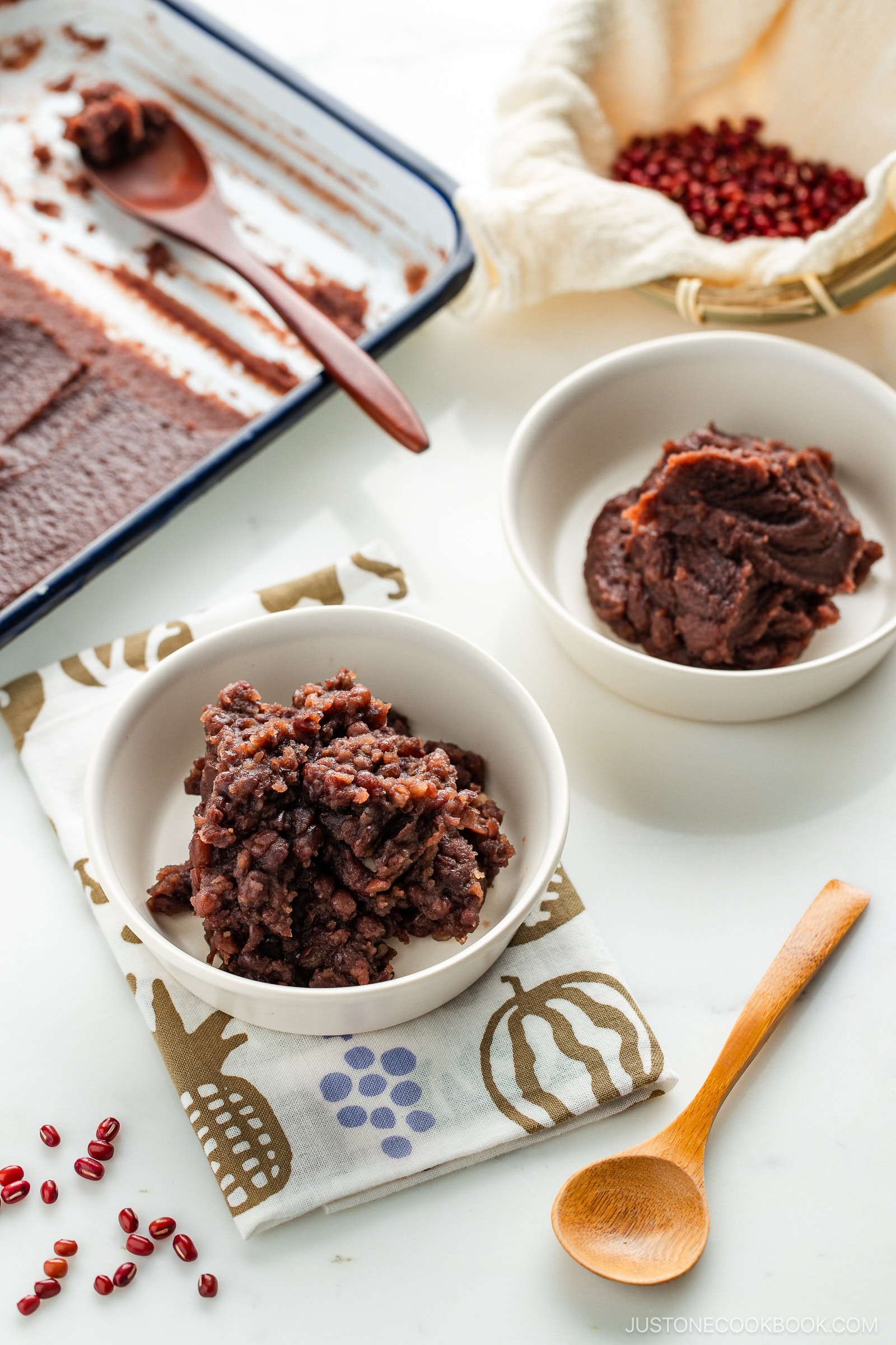 Two white bowls filled with different textures of sweet red bean paste sit on a patterned cloth; one paste is smooth, the other