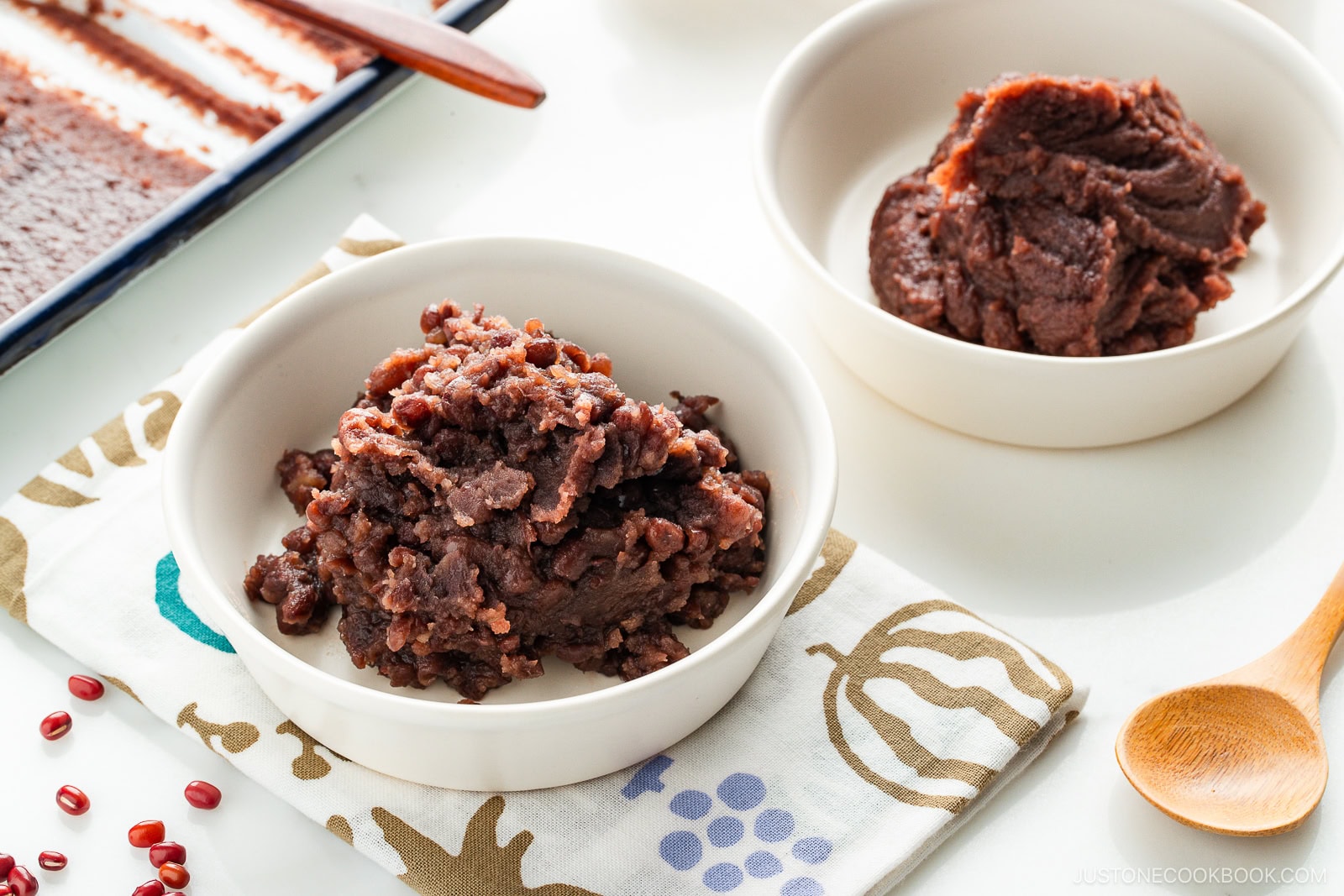 Two white bowls filled with sweet red bean paste sit on a patterned cloth; a wooden spoon and tray of spread red bean paste are beside them on a white surface.