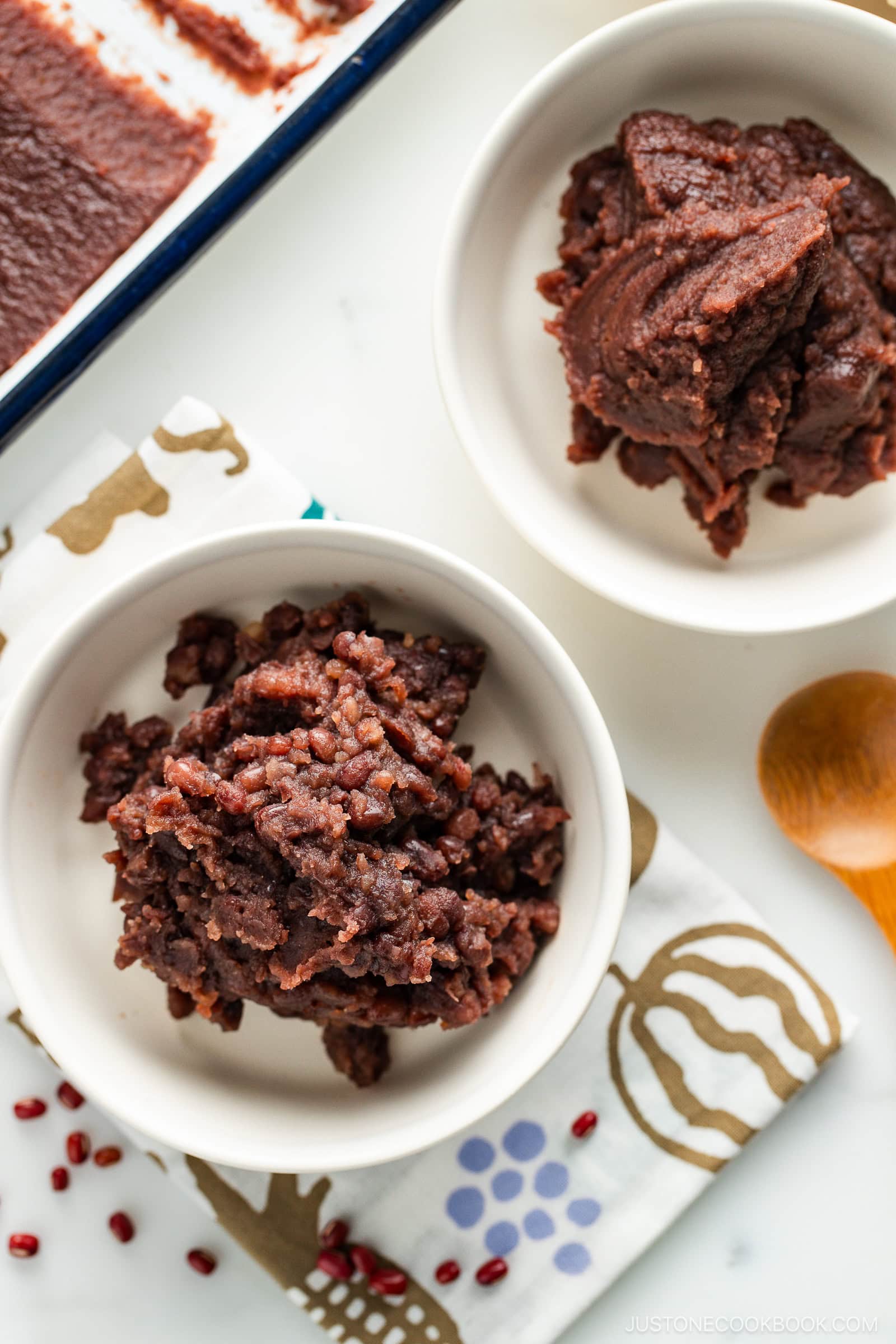 Two white bowls filled with different textures of sweet red bean paste sit on a patterned cloth; one paste is smooth, the other chunky, with a tray of more paste and a wooden spoon nearby.