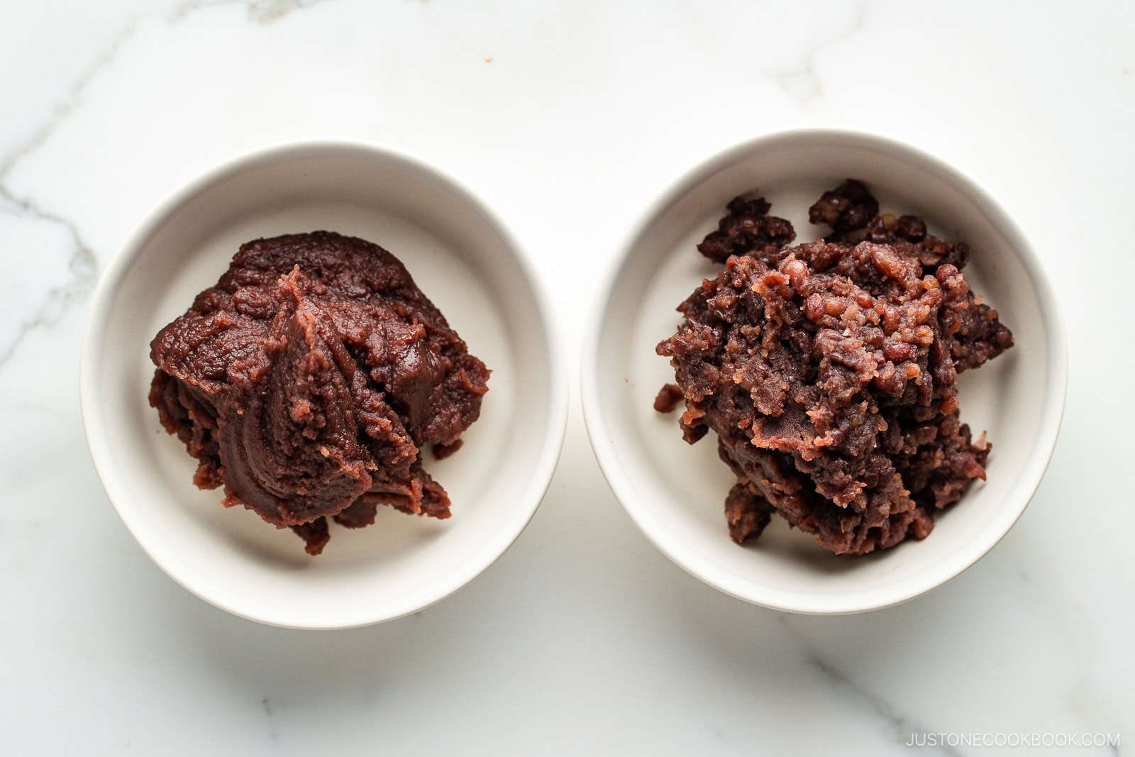 Two small white bowls on a marble surface, each containing a different type of chunky reddish-brown paste, likely varieties of sweet red bean paste (anko). The left paste is smoother, while the right paste is more textured.