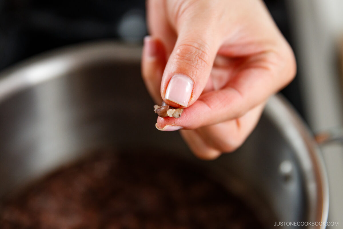 A close-up of a hand pinching a small amount of cooked beans above a pot, checking their doneness.
