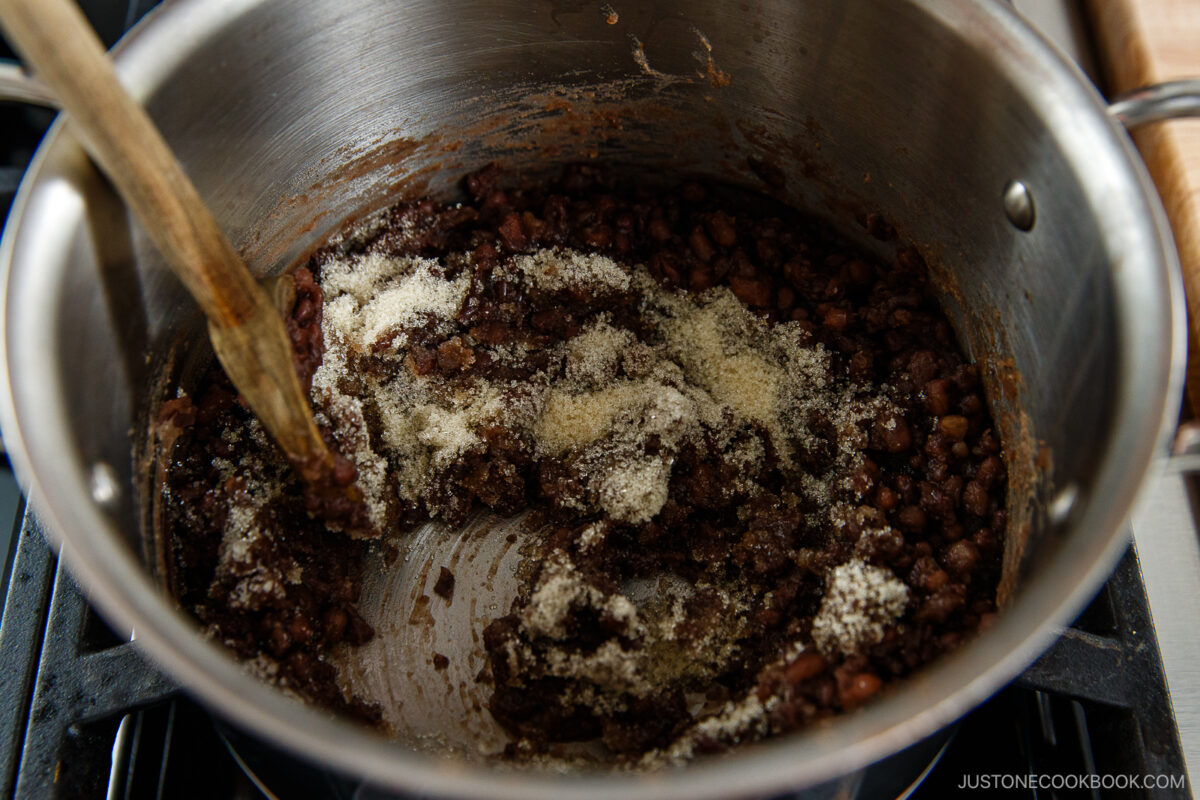 A pot of sweet red bean paste being stirred with a wooden spoon on a stove, with sugar sprinkled on top and partially mixed in.