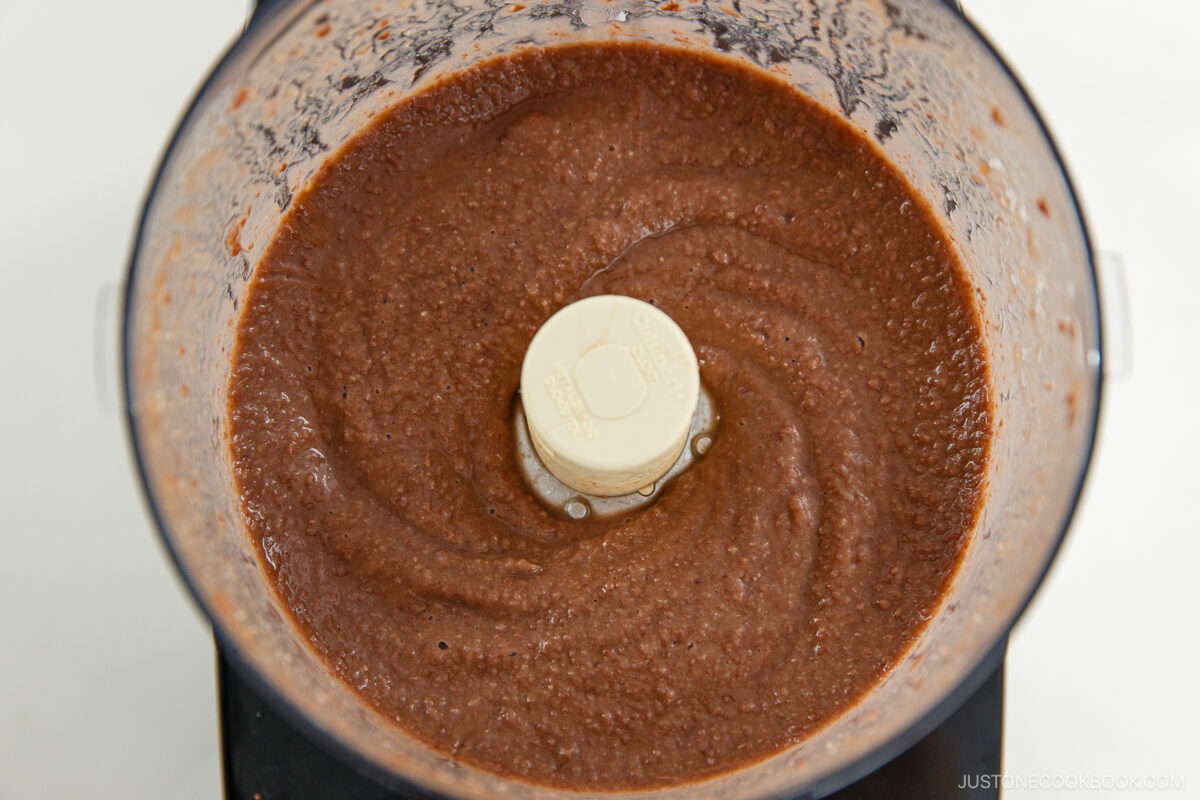 A food processor bowl filled with smooth, blended dark brown mixture, likely bean paste or batter, viewed from above.