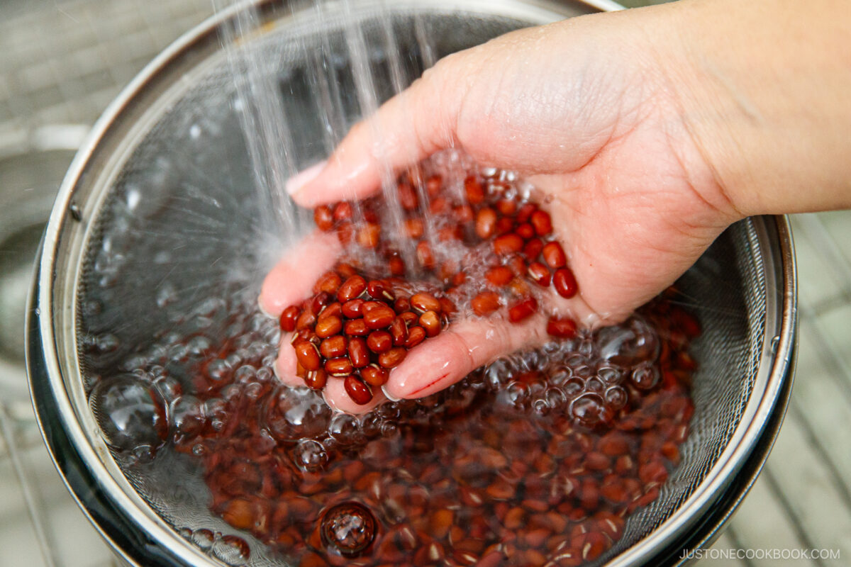 A hand holding red beans under running water, rinsing them in a metal strainer over a bowl.