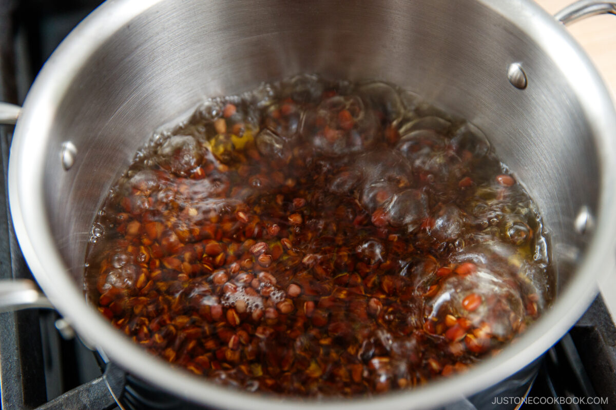 A stainless steel pot on a stove with small reddish seeds boiling in water, creating bubbling and steam on the surface.