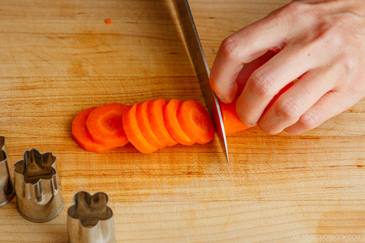 A hand slices a carrot into rounds on a wooden cutting board with a sharp knife. Metal vegetable cutters shaped like flowers are placed nearby.