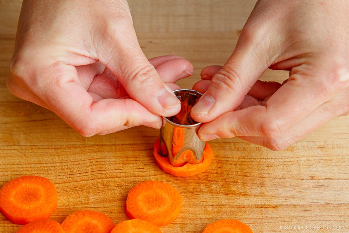 Hands pressing a metal flower-shaped cutter into a slice of carrot on a wooden cutting board, with several carrot slices nearby.