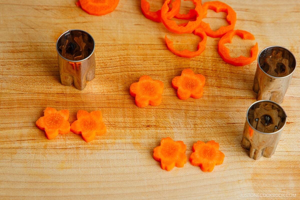 Sliced carrots cut into flower shapes on a wooden cutting board, with metal flower-shaped vegetable cutters beside them. Orange carrot rings and flower cutouts are arranged neatly.