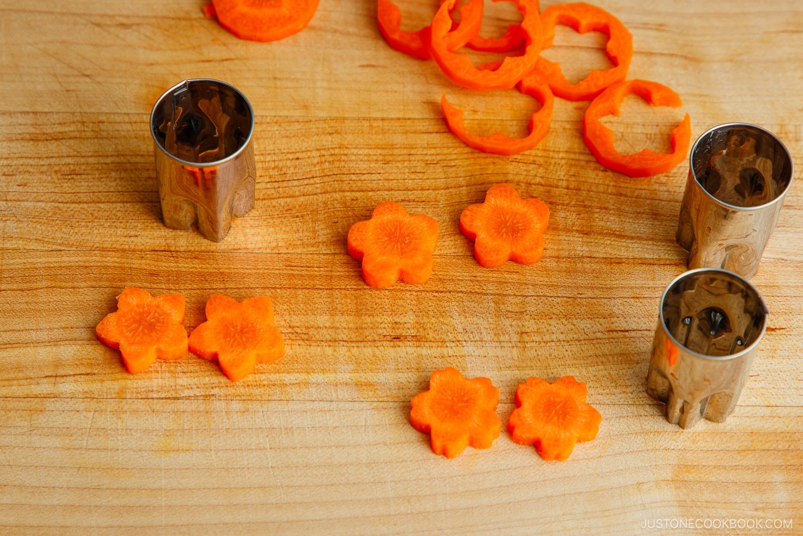 Sliced carrots cut into flower shapes on a wooden cutting board, with metal flower-shaped vegetable cutters beside them. Orange carrot rings and flower cutouts are arranged neatly.
