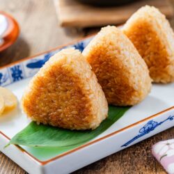 Three grilled Japanese rice balls (yaki onigiri) are arranged on a rectangular plate with a bamboo leaf, accompanied by pickled vegetables and a slice of pickled radish.
