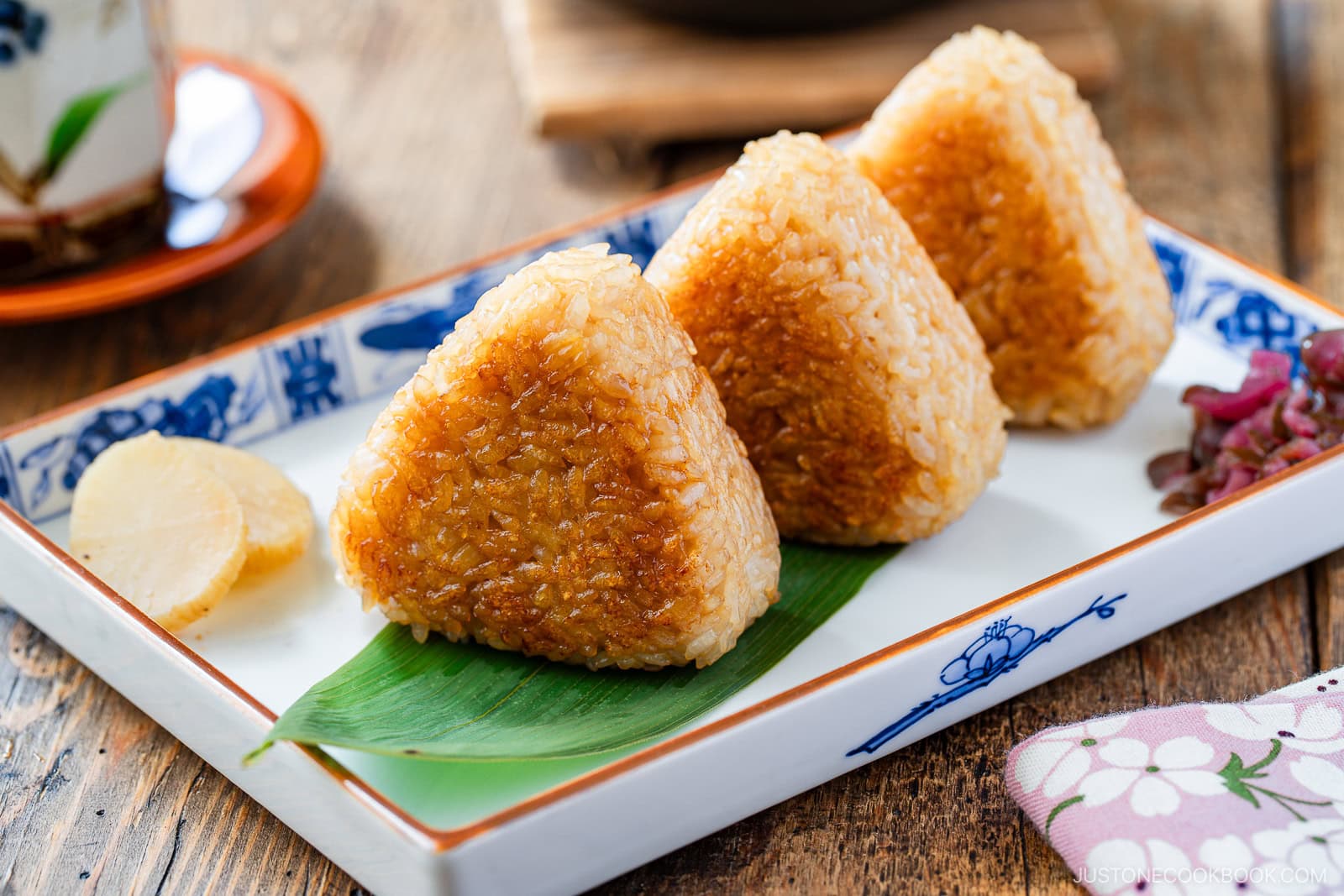 Three grilled Japanese rice balls (yaki onigiri) are arranged on a rectangular plate with a bamboo leaf, accompanied by pickled vegetables and a slice of pickled radish.