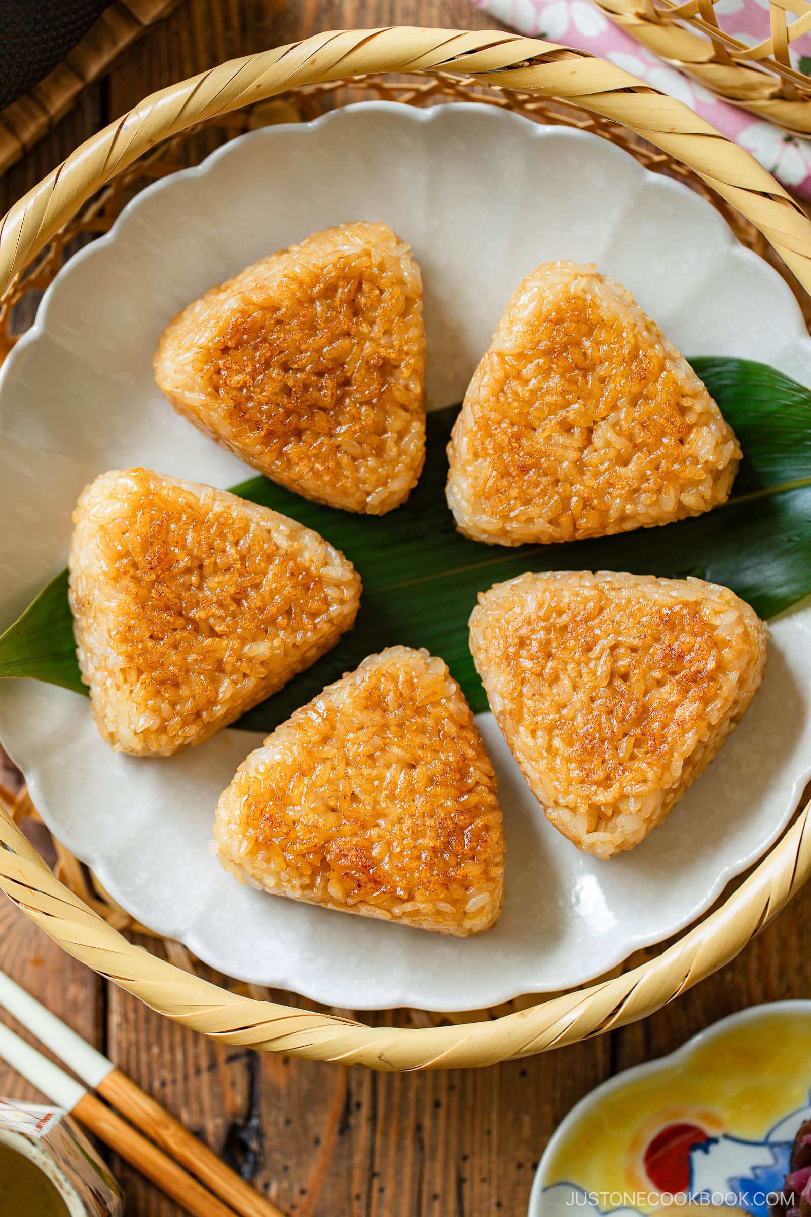 A plate with five golden-brown, grilled triangular rice balls (yaki onigiri) arranged neatly on a green leaf, set in a bamboo basket with chopsticks and a dipping sauce on the side.