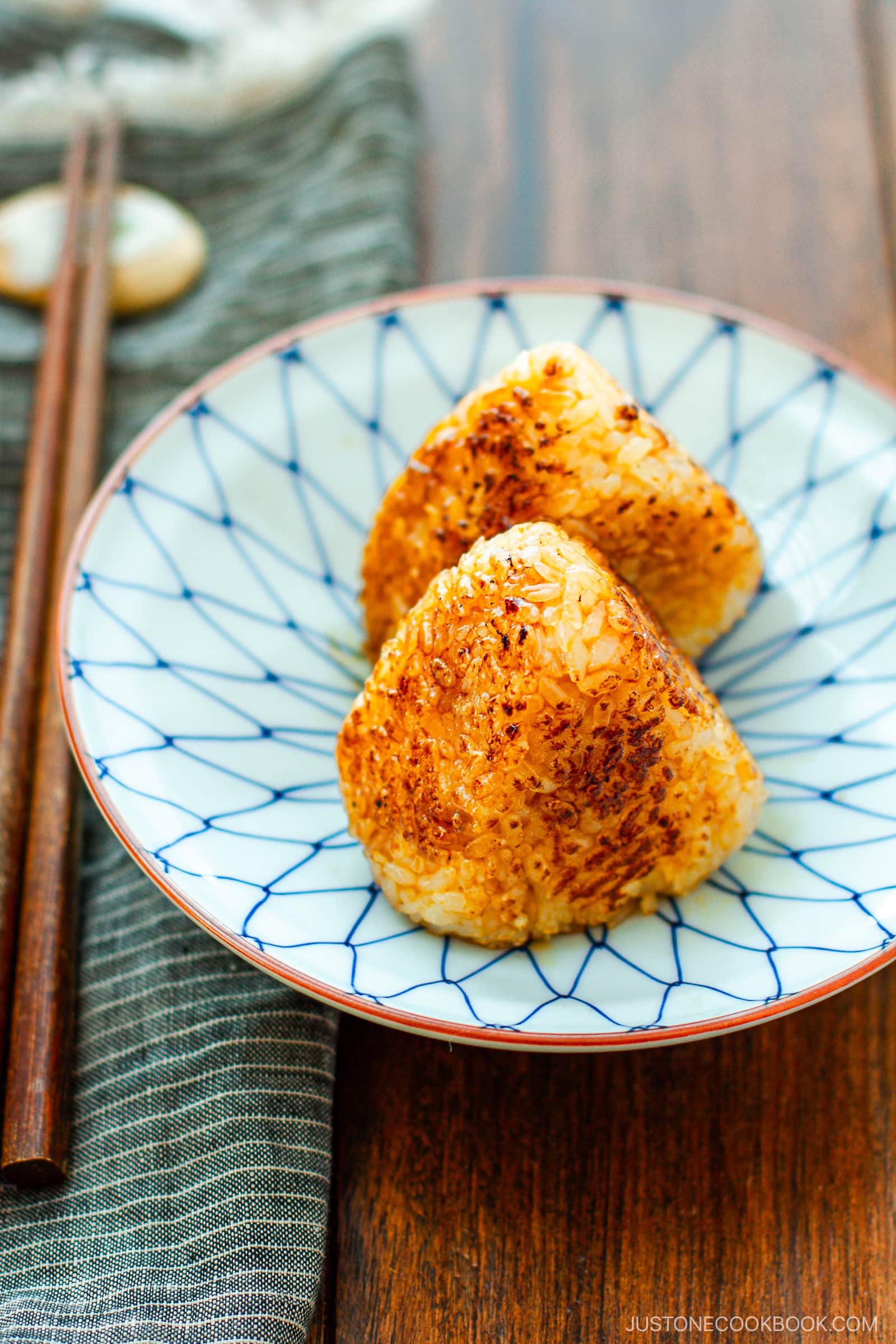 Two grilled Japanese rice balls (yaki onigiri) on a patterned blue and white plate, set on a wooden table with chopsticks and a striped napkin nearby.