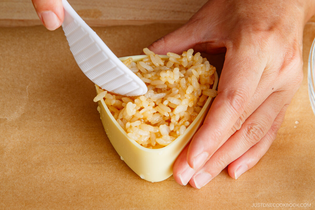 A close-up of hands pressing cooked rice into a yellow triangular mold using a white spatula, on a wooden surface.