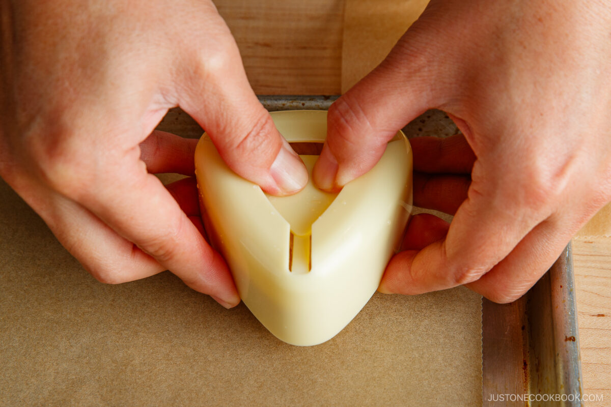 Two hands press a plastic heart-shaped mold onto parchment paper, shaping food inside the mold on a wooden surface.