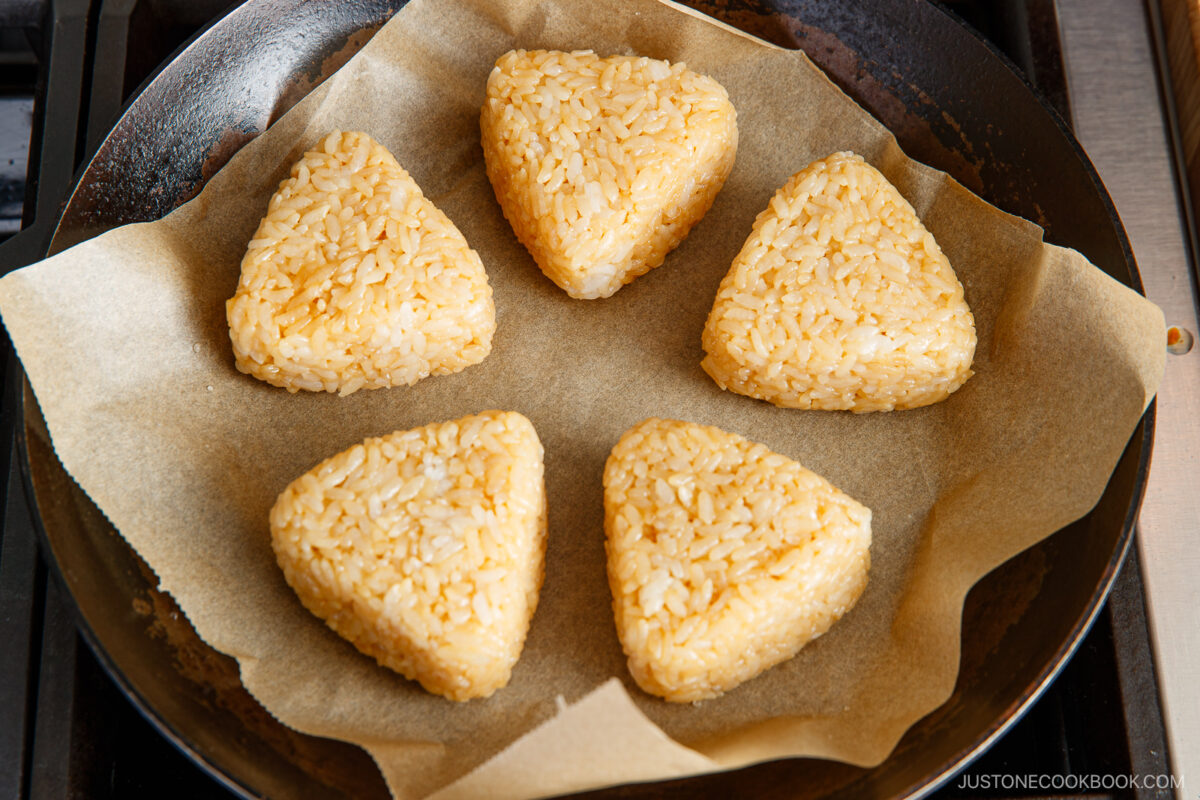 Five triangular rice balls are arranged in a circle on parchment paper in a skillet, ready to be cooked.
