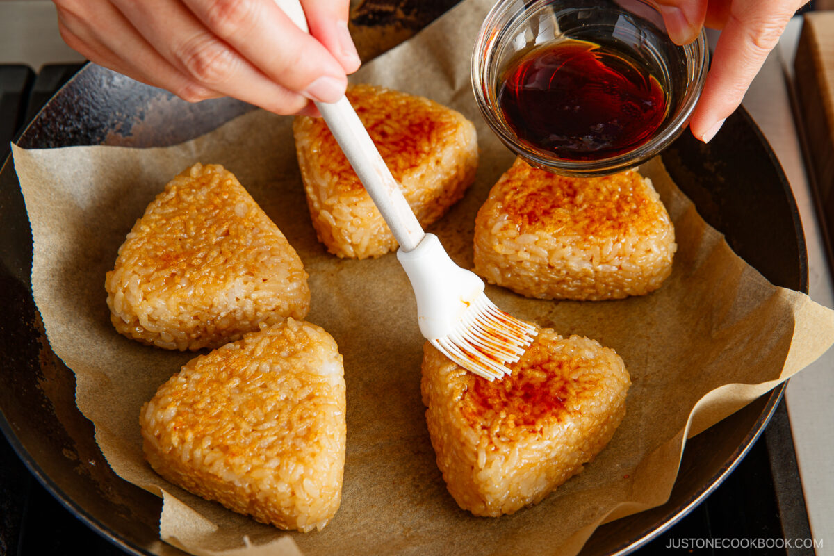 A hand uses a brush to apply soy sauce from a small bowl onto grilled rice balls, known as yaki onigiri, which are cooking on parchment paper in a pan.