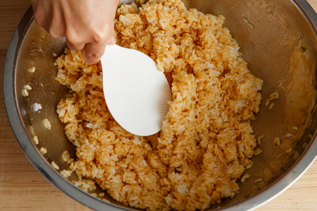 A hand mixes seasoned rice with a white rice paddle in a large metal bowl on a wooden surface.
