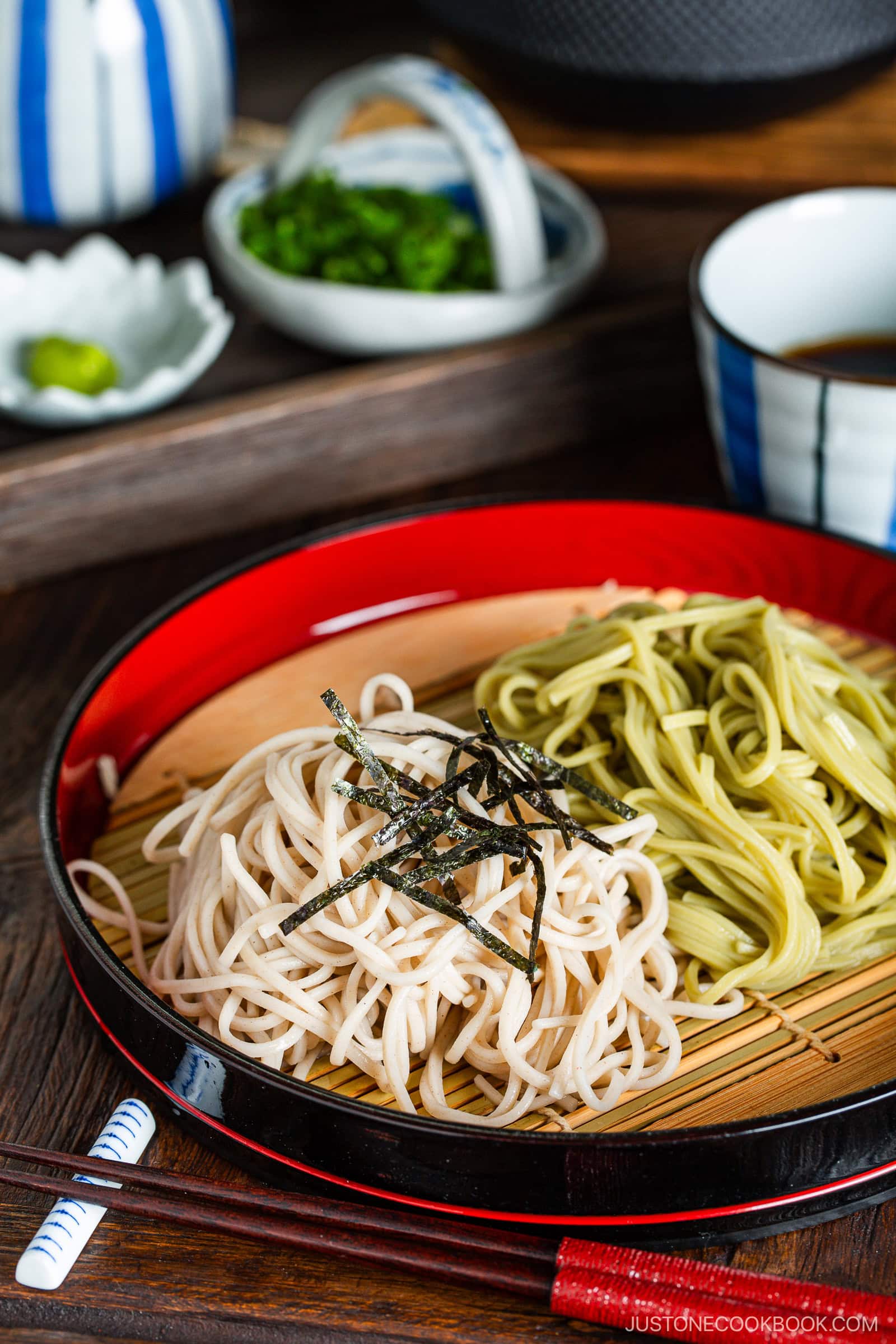 Zaru soba and zaru cha soba (cold buckwheat noodles and green tea noodles) served on a bamboo mat with shredded nori on top. Dipping sauce, green onions, and wasabi are in the background.