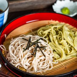 A red tray with bamboo mat holding two types of cold noodles, one white and one green, garnished with shredded seaweed, served with dipping sauce and condiments in bowls nearby. Chopsticks rest on the side.