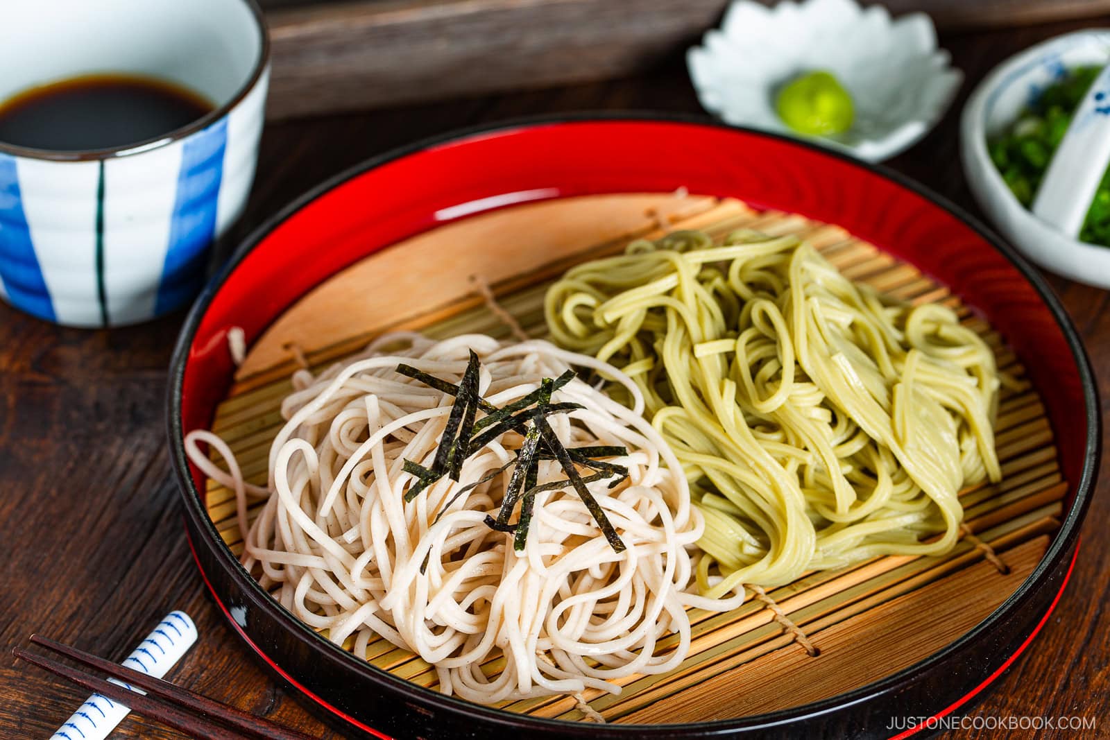 A red tray with bamboo mat holding two types of cold noodles, one white and one green, garnished with shredded seaweed, served with dipping sauce and condiments in bowls nearby. Chopsticks rest on the side.