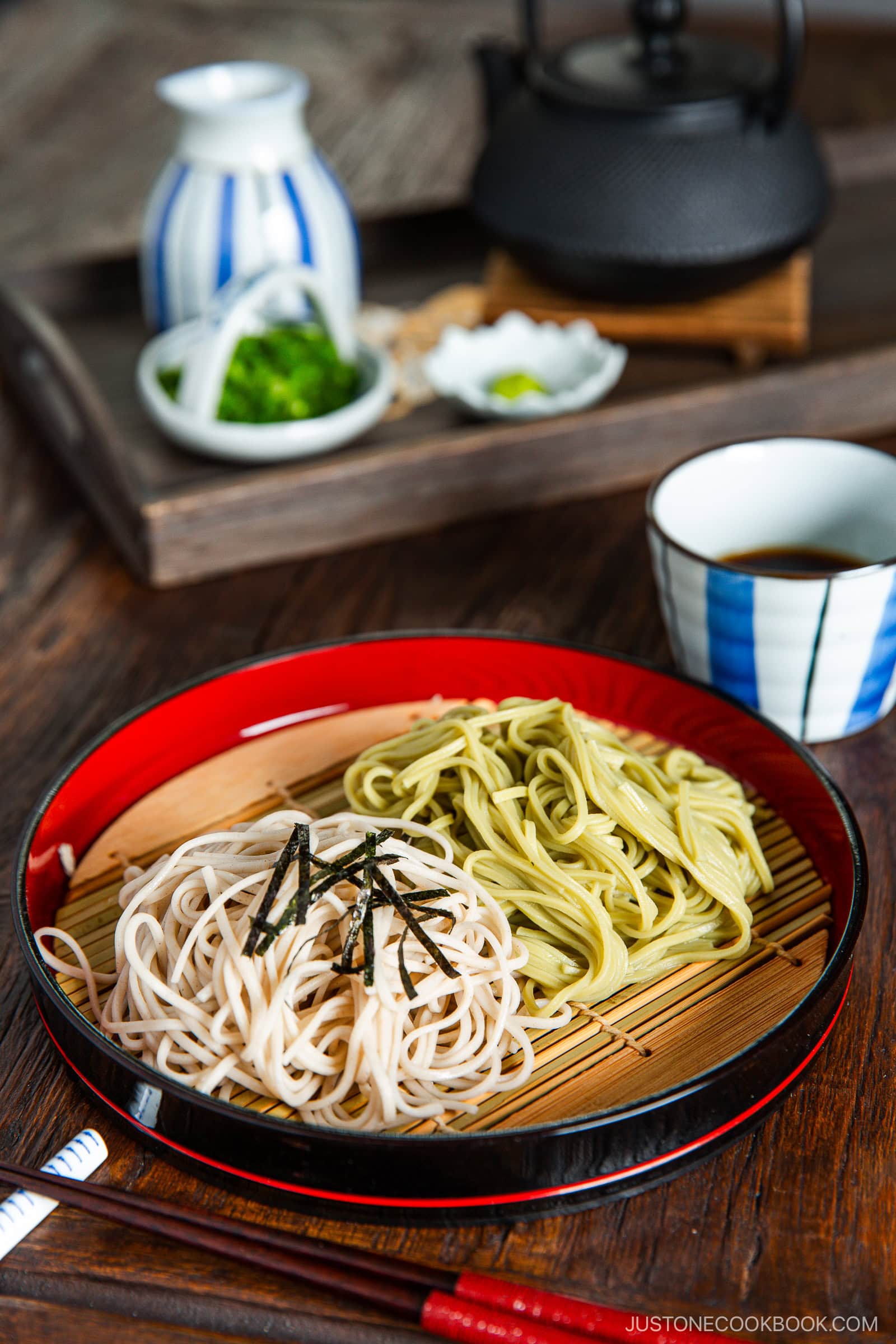 A plate of cold soba and green tea noodles garnished with shredded seaweed, served on a bamboo mat with dipping sauce, tea, and condiments in the background.