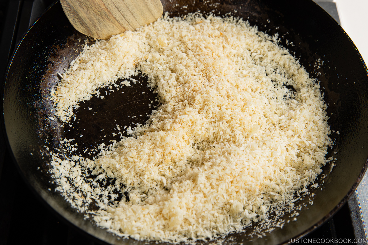 A close-up of panko breadcrumbs being toasted in a dark frying pan, stirred with a wooden spatula. The breadcrumbs are turning golden brown.