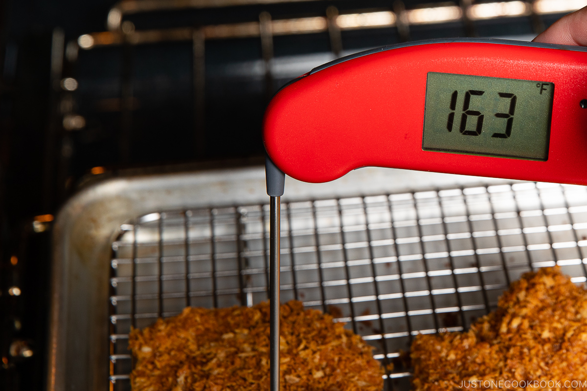 A red digital meat thermometer reads 163°F while inserted into a breaded piece of meat on a wire rack over a baking tray inside an oven.