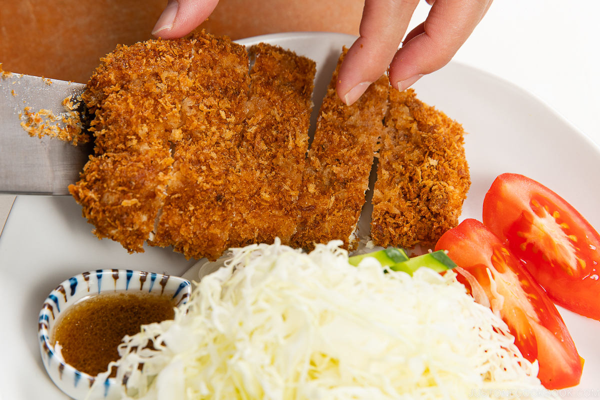 A hand uses a knife to slice golden, crispy breaded cutlet on a plate with shredded cabbage, tomato wedges, cucumber slices, and a small dish of dipping sauce.