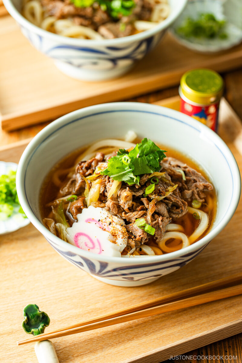 A bowl of udon noodle soup topped with sliced beef, fish cake, green onions, and cilantro, set on a wooden table with chopsticks and a condiment container nearby.