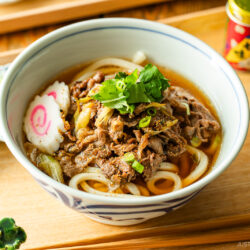 A bowl of beef udon with sliced beef, green onions, narutomaki (fish cake), and fresh herbs in broth, served on a wooden tray with chopped green onions and a bottle of seasoning on the side.