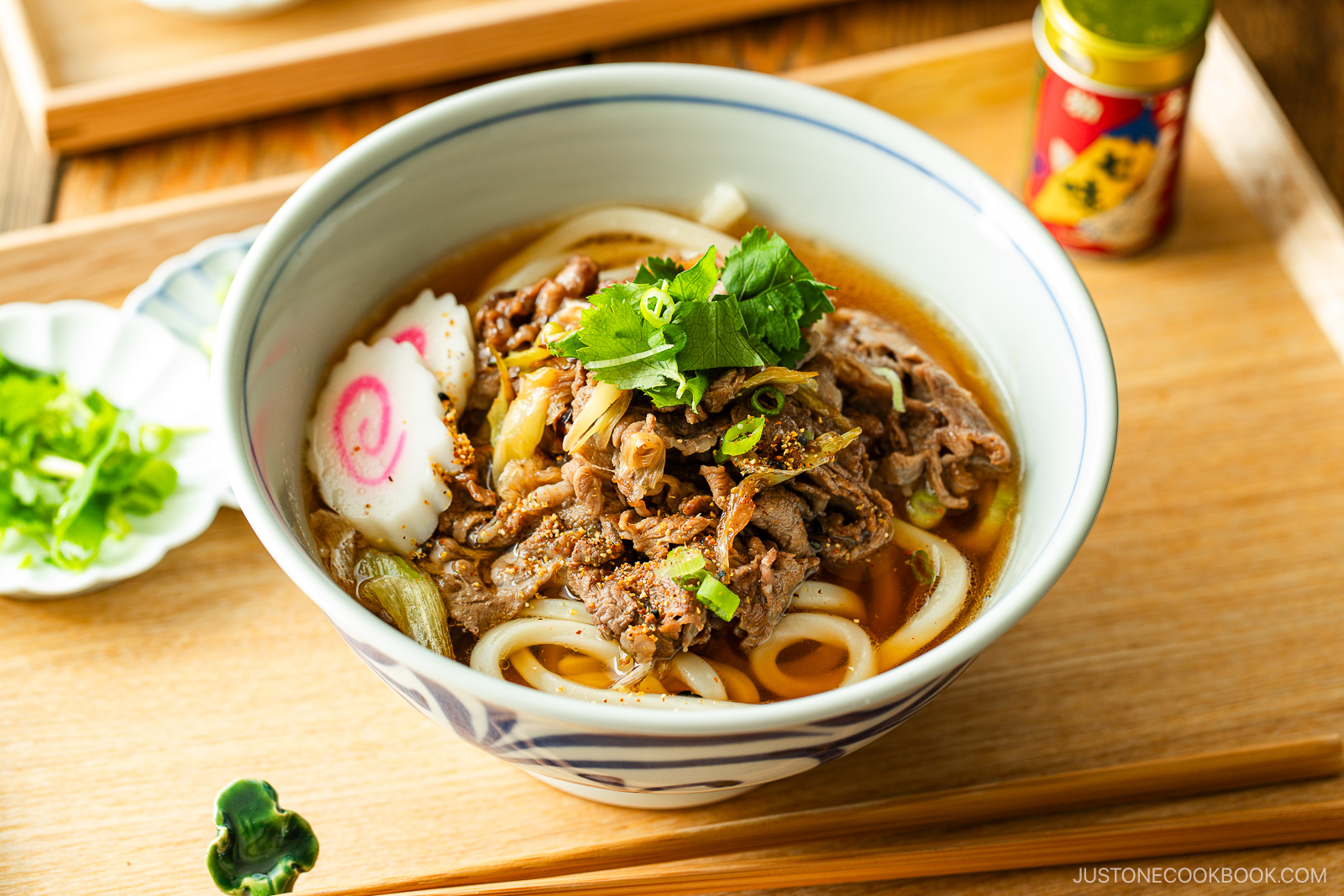 A bowl of beef udon with sliced beef, green onions, narutomaki (fish cake), and fresh herbs in broth, served on a wooden tray with chopped green onions and a bottle of seasoning on the side.