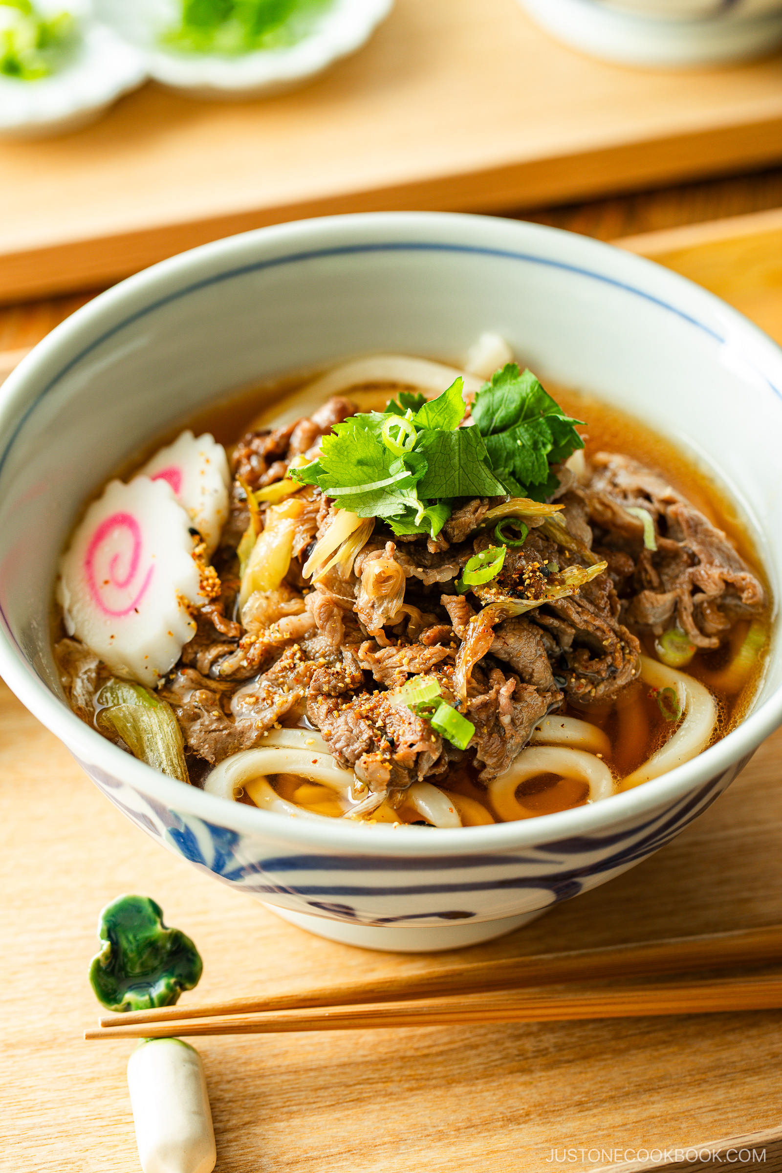A bowl of Japanese udon noodle soup topped with sliced beef, fish cake, green onions, and fresh herbs, with chopsticks and a chopstick rest on the wooden table beside it.