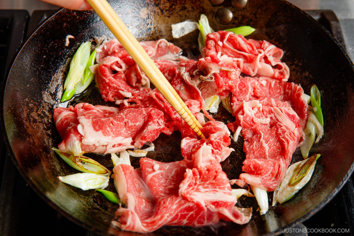Thin slices of raw beef and chopped green onions being stir-fried in a hot pan with a pair of chopsticks.