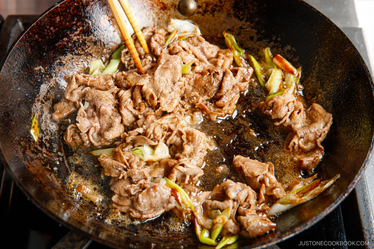 Thin slices of beef and pieces of green onion are being stir-fried in a sizzling, oily pan with wooden chopsticks on a stovetop.