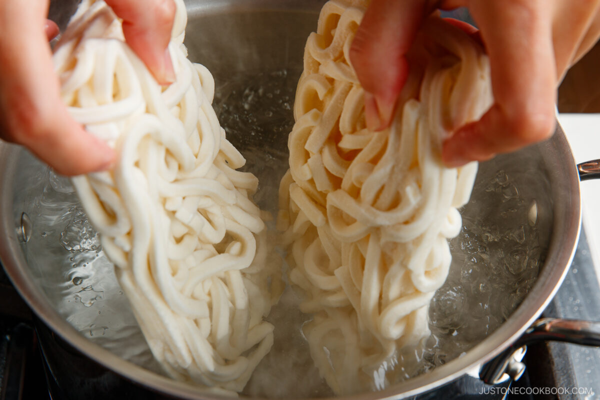 Two hands place blocks of uncooked udon noodles into a pot of boiling water on a stovetop.