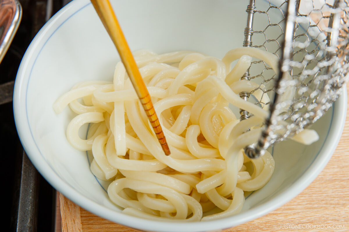 A bowl of cooked udon noodles being placed into the bowl with chopsticks and a metal strainer.