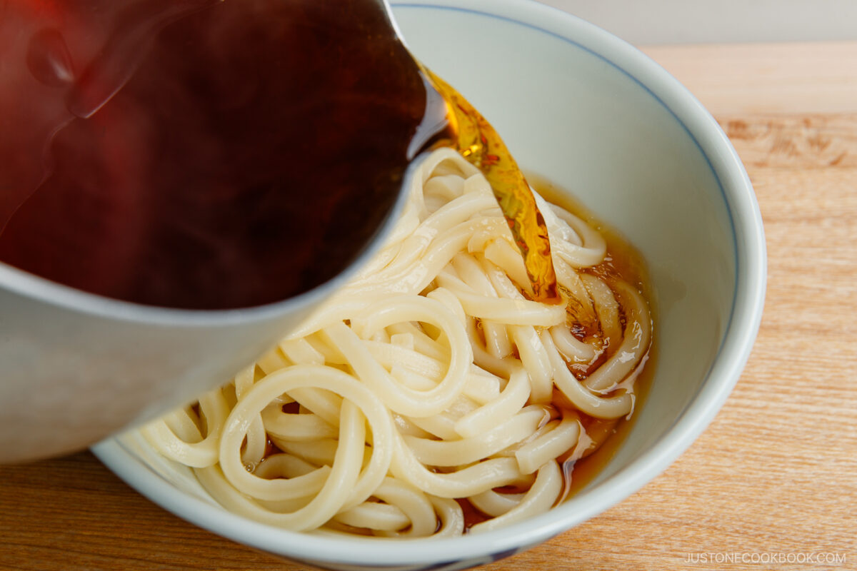 A bowl of udon noodles is being filled with a dark, savory broth poured from a pot, set on a light wooden surface.