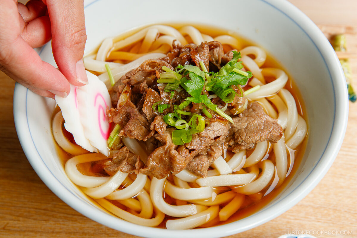 A hand places a slice of narutomaki fish cake into a bowl of udon noodle soup topped with sliced beef and chopped green onions.