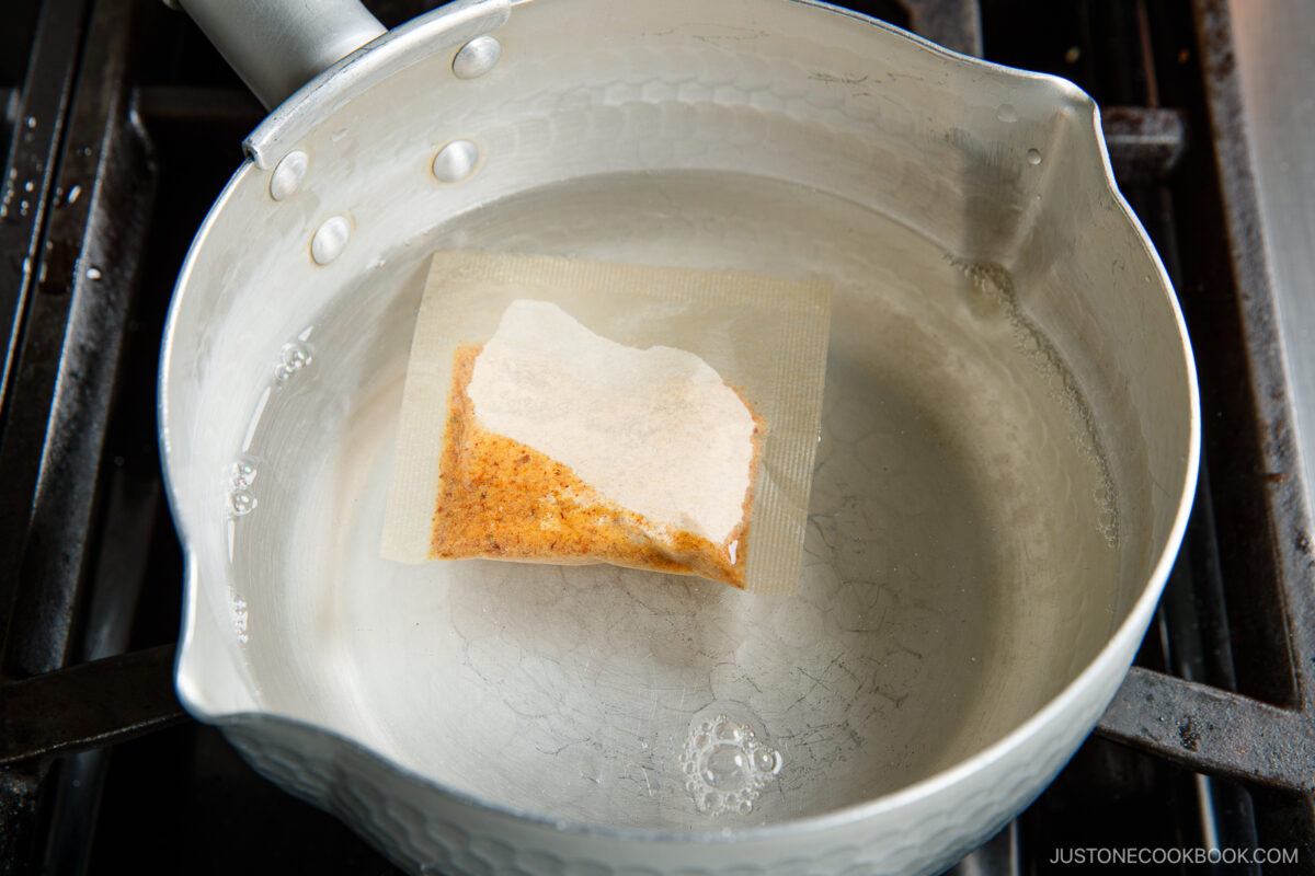 A small mesh bag filled with seasoning floats in a saucepan of water on a stovetop, as the water begins to heat but has not yet reached a boil.